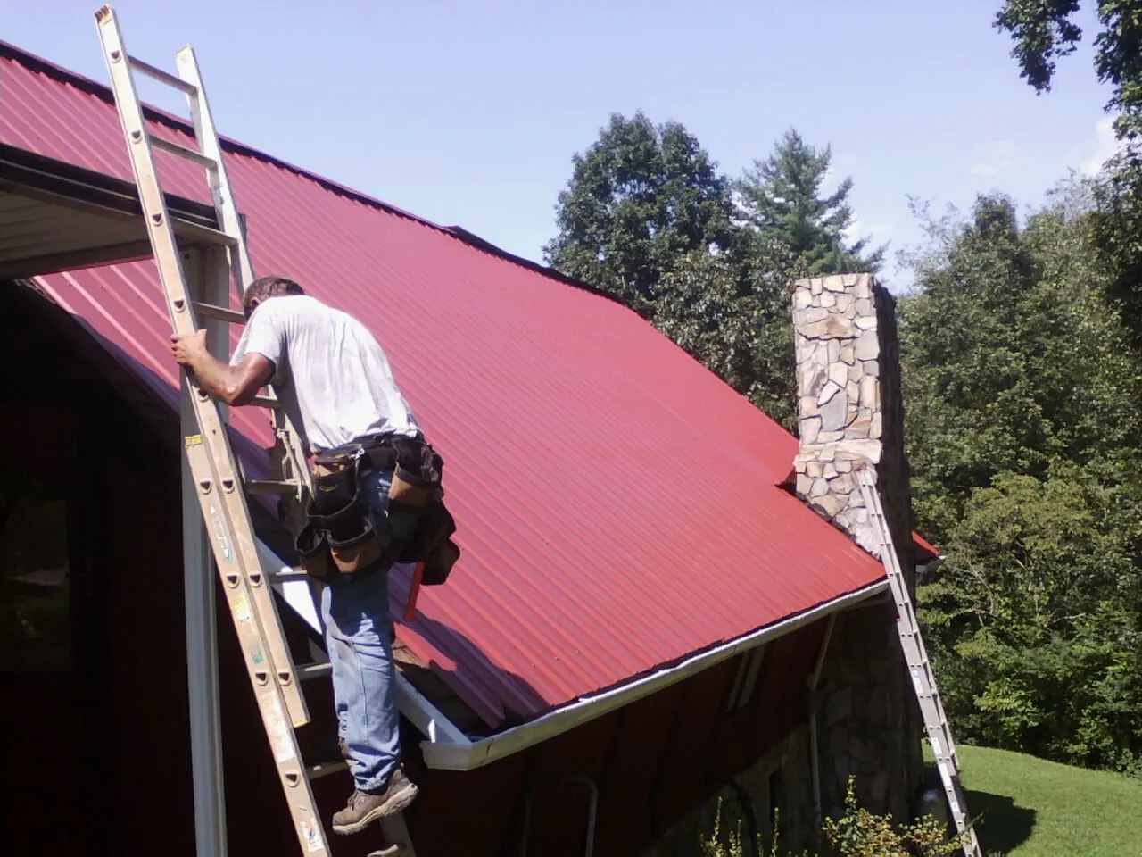 Roofer on a ladder beside a red metal roof and stone chimney — Soffit & Fascia Repair by Roofing Brothers in Stewartsville